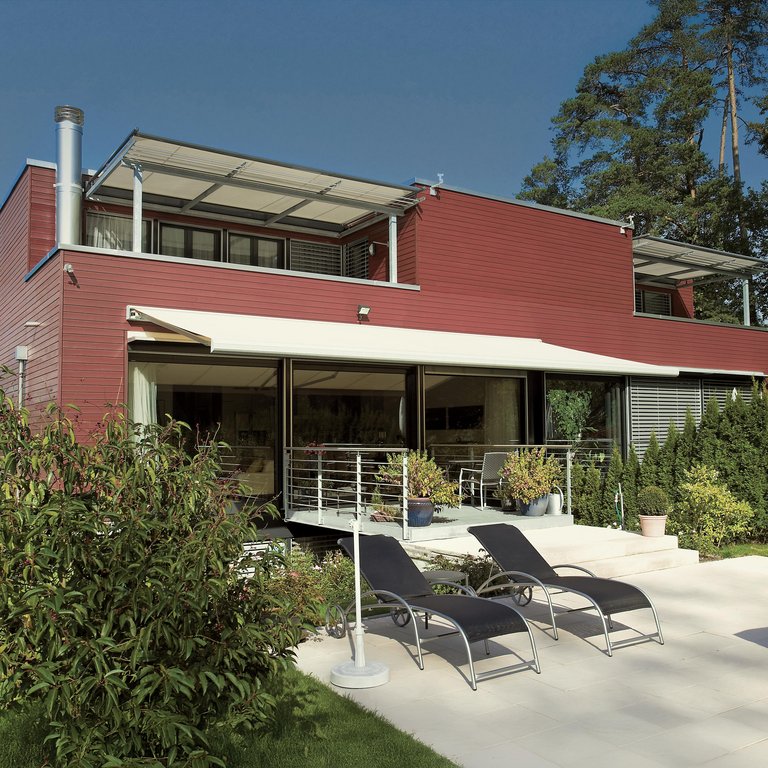Maison moderne avec terrasse, pergola et chaises longues, entourée de verdure et d’un ciel bleu.