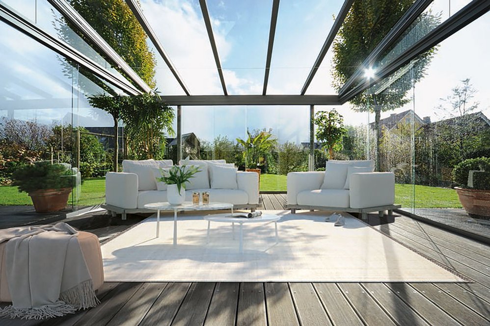 Intérieur d'un jardin d'hiver avec canapés blancs et plante verte sur un sol en bois, lumineux et ouvert sur l'extérieur.