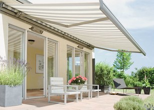 Vues de l'extérieur d'une maison avec une grande marquise rayée, des sièges et une végétation en pot. 