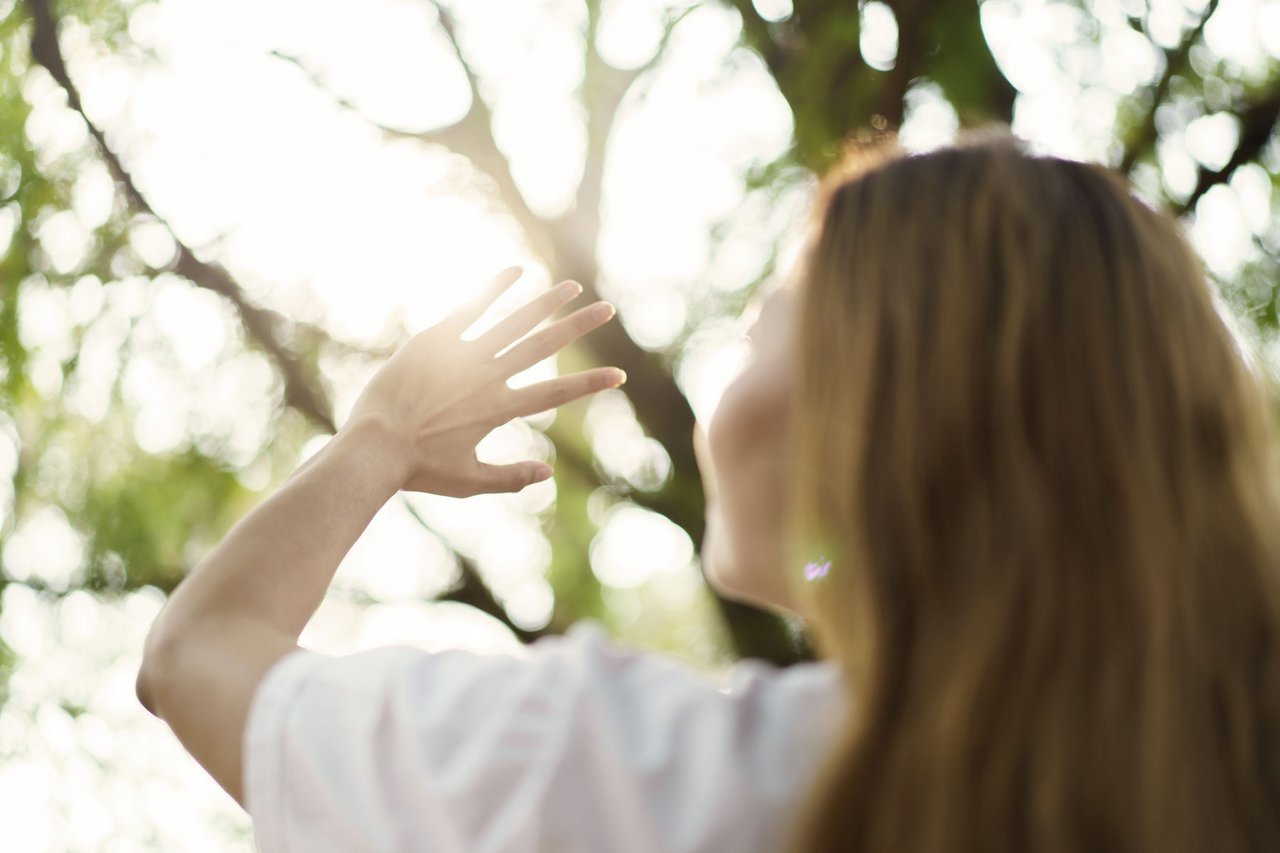 Eine Person mit einer Hand in der Luft, im Hintergrund Sonnenstrahlen und grüne Blätter.