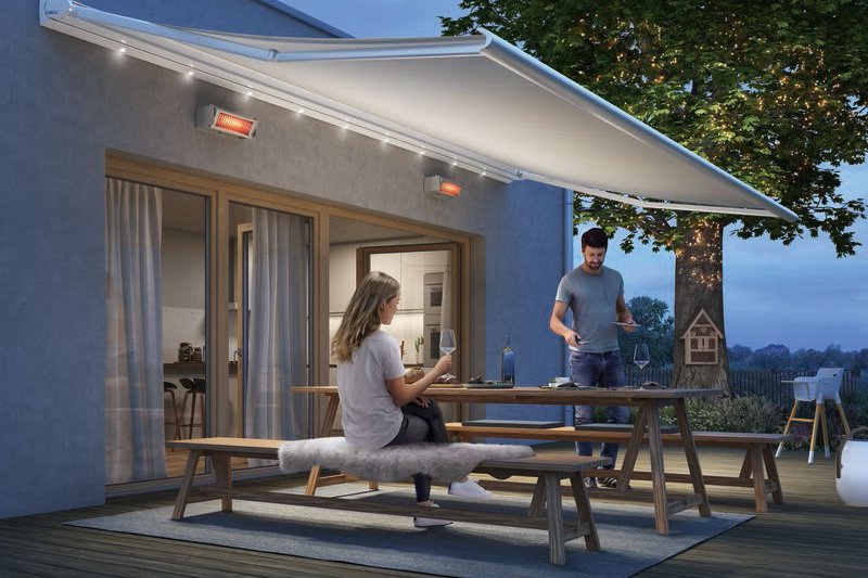 Scène de vie en extérieur avec des personnes, une terrasse éclairée et une marque de protection solaire déployée. Scène de vie en extérieur avec des personnes, une terrasse éclairée et une marque de protection solaire déployée.