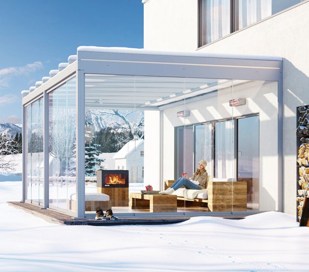Terrasse avec une structure en verre, des meubles en bois et un poêle à bois sous un ciel ensoleillé. 