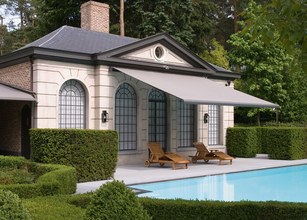 Maison en pierre avec une terrasse, un auvent gris, et une piscine entourée de verdure. 