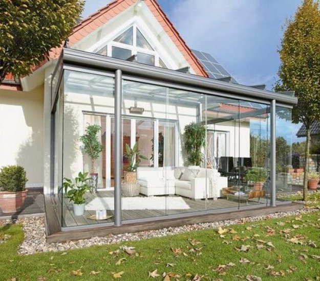 Terrasse avec une construction en verre, intégrée à une maison, entourée de verdure et de mobilier intérieur. 