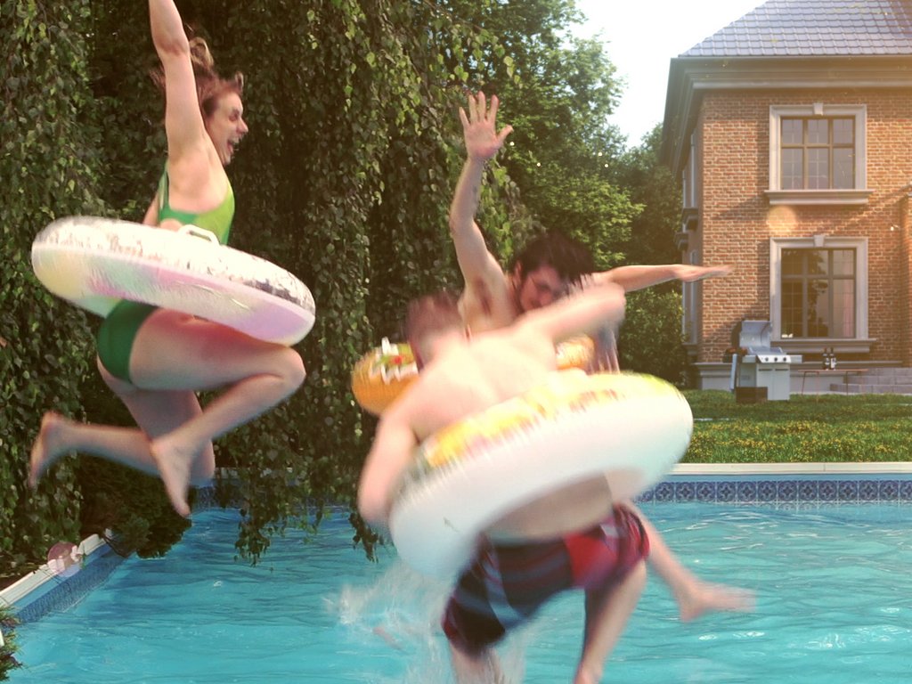 Trois personnes sautant dans une piscine, avec des bouées colorées. Ambiance estivale et joyeuse.