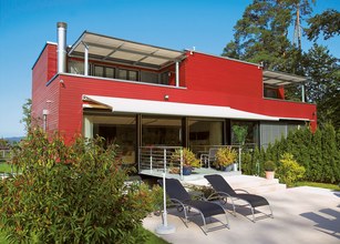 Maison moderne avec façade rouge, terrasse et chaises longues, entourée de végétation. 