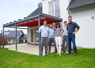 Groupe de personnes souriantes sous une structure de terrasse avec une toiture rouge et un arrière-plan verdoyant. 