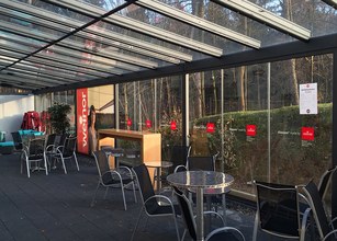 Espace extérieur couvert avec des chaises et tables, parois en verre et une végétation à l'arrière-plan. 