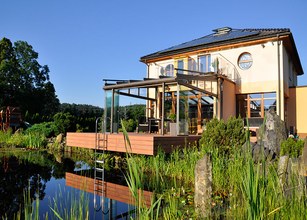 Maison moderne avec terrasse en bois, véranda en verre, entourée de verdure et d'eau. Ciel bleu clair. 