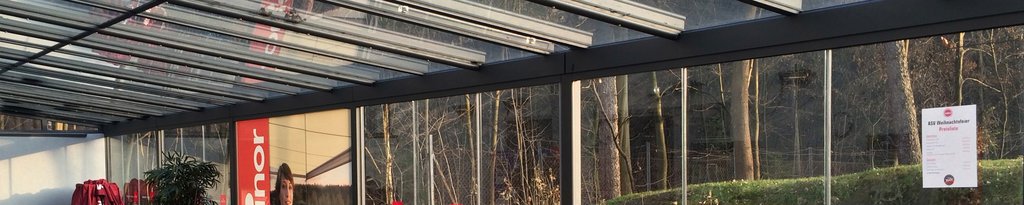 Vue d'une structure avec des murs en verre et un toit en lames orientables, en métal gris.