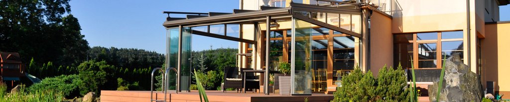 Vue d'une terrasse avec une structure en verre et bois, entourée de verdure et de ciel bleu.