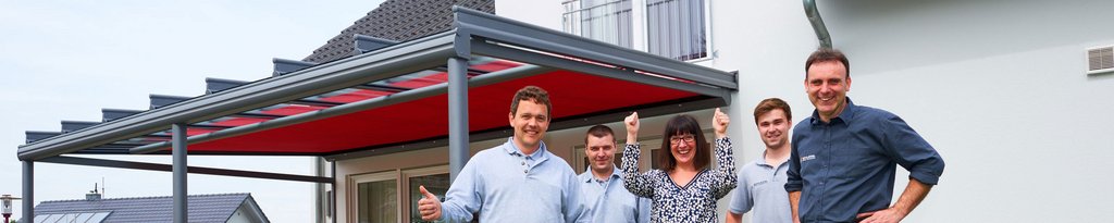 Équipe souriante sous une structure de toiture rouge, avec une maison blanche en arrière-plan.