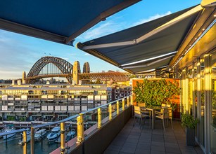 Vue d'une terrasse avec une ombre en tissu, un pont et des bâtiments en arrière-plan sous un ciel bleu. 