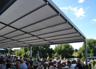 Une grande structure de toit en gris avec des personnes assises en dessous, entourées de verdure et d'eau. 